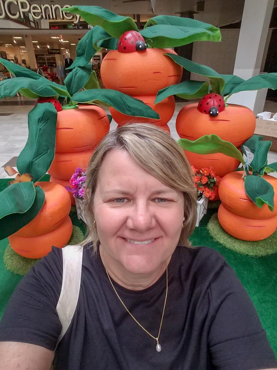 Mary taking a selfie in front of large orange carrots with lady bugs on them with JC Penny in the background.